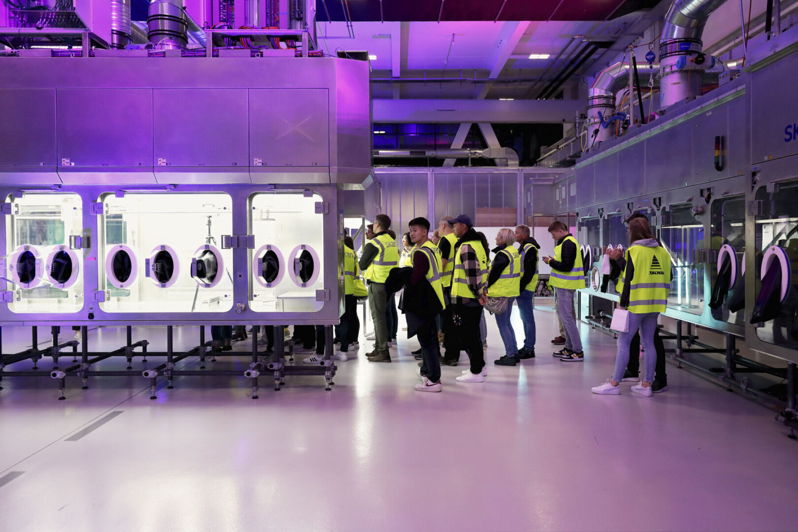 Besucher in gelben Warnwesten besichtigen eine Industrieanlage mit grossen sterilen Boxen.