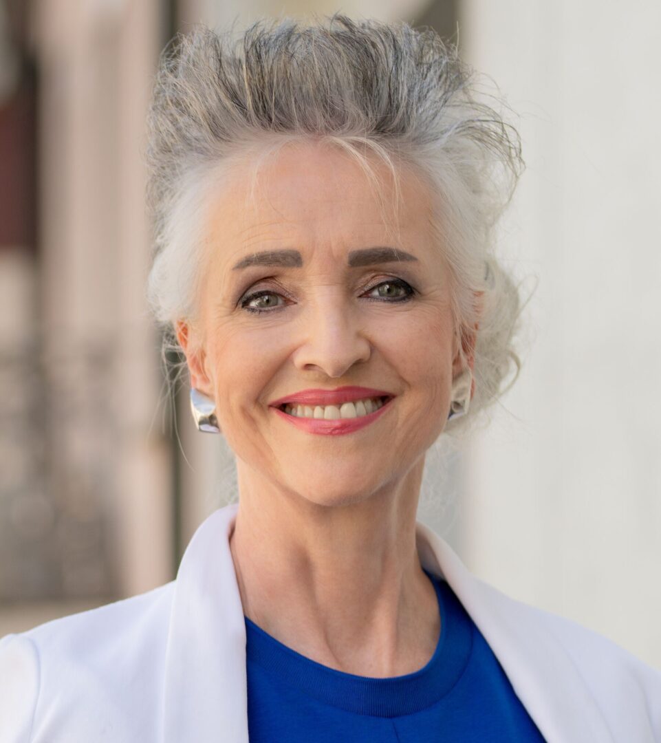Carmen Walker Späh Porträt einer lächelnden Frau mit grauem, hochgestyltem Haar in weissem Blazer und blauem Shirt.