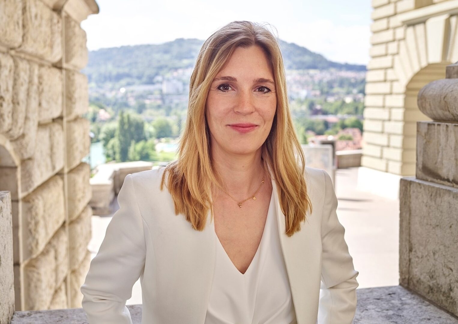 Corina Gredig Porträt einer lächelnden Frau mit blonden Haaren in einem weissen Blazer vor einer Stadtkulisse.