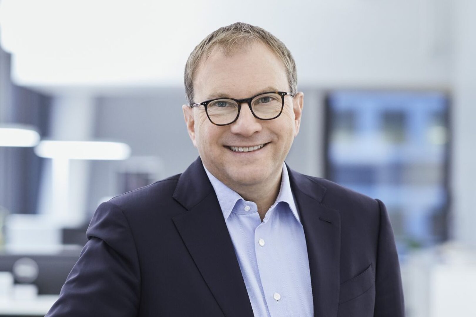 Martin Hirzel Porträt eines lächelnden Mannes mit Brille in einem dunklen Sakko und hellem Hemd.