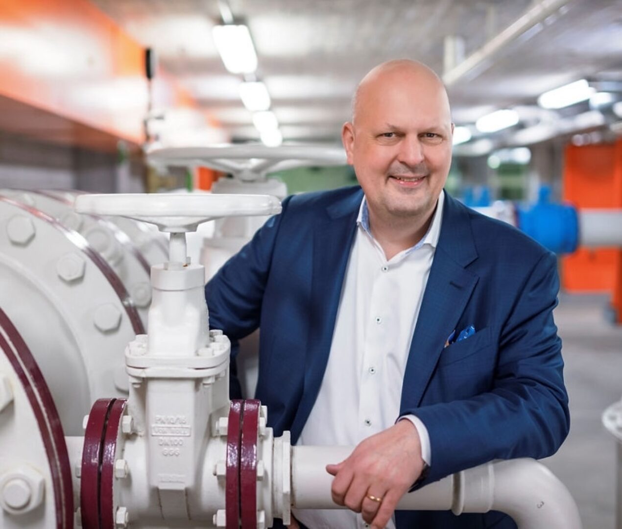 Michael Baumer Lächelnder Mann im blauen Sakko lehnt an einer grossen weissen Industrieanlage mit Ventilen.
