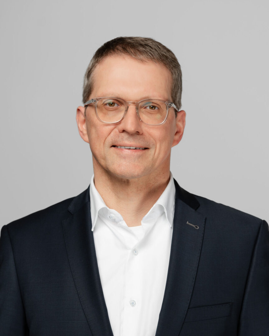 Patrik Meli Porträt eines lächelnden Mannes mit Brille, weissem Hemd und dunkelblauem Sakko vor grauem Hintergrund.