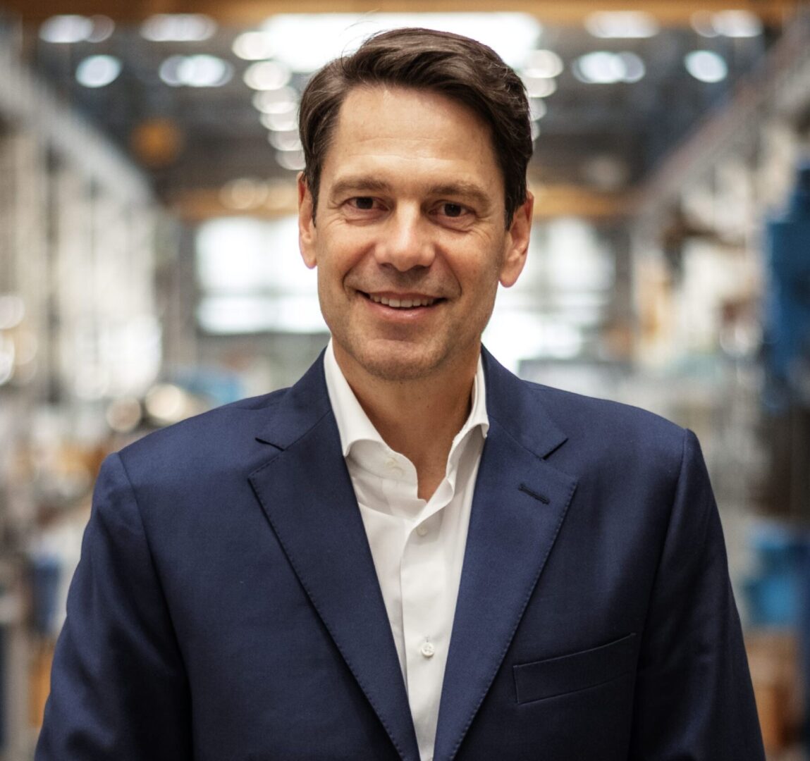 Dr. Stefan Brupbacher Porträt eines lächelnden Mannes in dunkelblauem Sakko und weissem Hemd vor industriellem Hintergrund.
