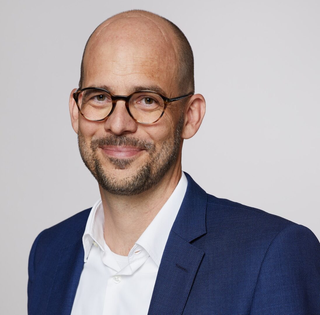 Yves Bisang Porträt eines glatzköpfigen Mannes mit Brille, Bart, weissem Hemd und blauem Sakko.