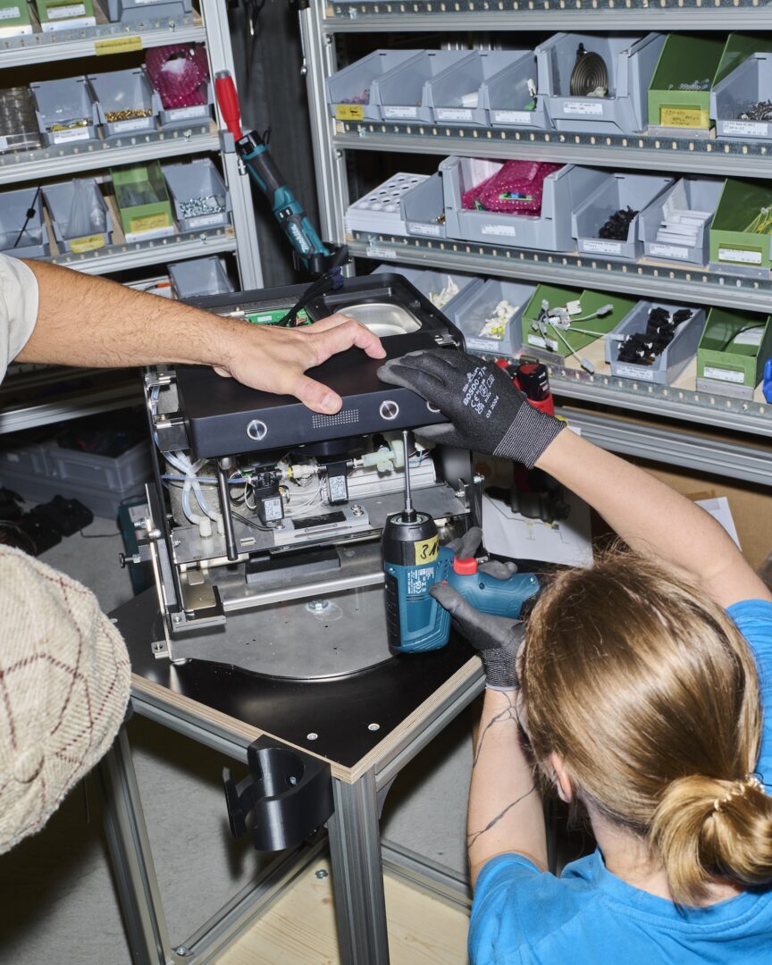 INZH27 ZURIGA Florian Spring 02 Two people assembling a technical device with a power drill in a workshop.