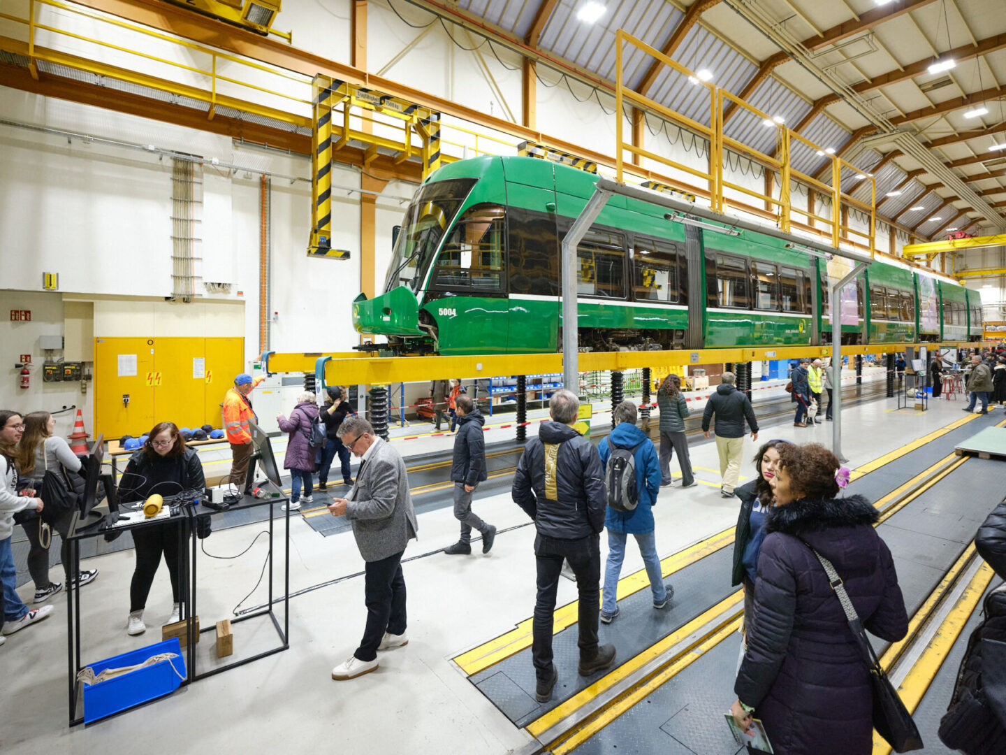 INRB24 Foto Samuel Bramley klein 103 Menschen besichtigen eine grüne Tram, die in einer Werkstatt auf einer Hebebühne steht.